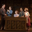 Three men and two women sitting behind a piano talking in front of a brown background