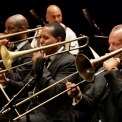 Three men, wearing black suits with black and white dotted ties, are playing the trombone sitted in a line. Drum player is visible in the background.