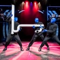 3 bald men dressed in black holding a piece of large white pipe using it as a drum