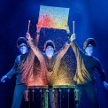 Three bald blue men dressed in black playing drums in front of a black background. Colored paint is splashing off the drums. Two of them are holding up a canvas with paint splattered on it.