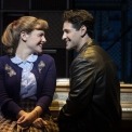 Man in a black leather jacket and woman in a purple cardigan sitting at a piano and smiling at each other