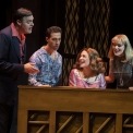 Two men and two women sitting behind a piano singing in front of a brown background