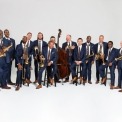 15 men, each wearing a blue suit, brown tie, and brown shoes, with a crisp white background. All men, except one, are holding a brass, wind, or percussion instrument. All of the men are smiling, some laughing.