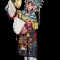 A Chinese woman wearing a headdress and robe made of ornate red, white, yellow, and blue fabric stands holding a gold fan with flowers on it at an angle near her face in front of a black background.