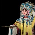 A Chinese woman wearing a headdress and robe made of ornate red, white, yellow, and blue fabric stands holding a gold fan parallel to the floor with her other hand up near her ear in front of a black background.