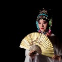A Chinese woman dressed in white with a gold and blue headband stands holding a gold fan directly in front of her chest in front of a black background.