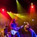 A man with long hair singing while playing guitar stands with two guitar players to his left, a long haired man leaning back with his arm in the air singing into a microphone and a man playing guitar to his left underneath red and green stage lights.