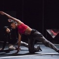 Woman in red top and grey pants on one hand and one knee reaching over her head with a person in black behind her mimicking her all in front of a black background.