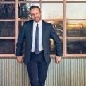 A man in a blue suit and tie and a white button down standing in front of a window and smiling at the camera.