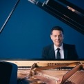 Michael Feinstein in a black suit and black tie and a white shirt sitting behind an open grand piano looking at the camera in front of a blue background.