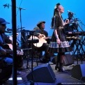 Woman wearing a black dress singing into a microphone while playing a percussion instrument with five other people in black playing instruments around her in front of a blue background.