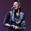 Woman wearing a black top singing into a microphone while playing a percussion instrument in front of a purple background.