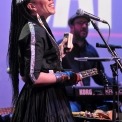 Woman wearing a black dress singing into a microphone while playing a percussion instrument and a man in black playing the keyboard behind her all in front of a purple background.