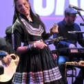 Woman wearing a black dress and smiling while playing a percussion instrument and a man in black playing the keyboard behind her all in front of a purple background.