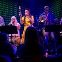 A woman in a yellow dress singing and holding an instrument with four background singers behind her all in front of a green background.