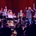 6 people singing and 3 people seen playing instruments behind them. Everyone is on a sage in front of a red background.