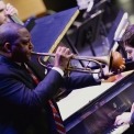 Male african american, wearing a black suit with a red, white, and blue striped tie, is playing a trumpet with a plunger. White man playing the piano to the trumpet player's left. And a male cello player is also visible behind the trumpet player.