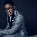 An African American man wearing glasses sits in front of a dark blue background wearing a gray suit.