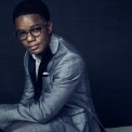 An African American man wearing glasses sits in front of a dark blue background wearing a gray suit.