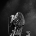 A black and white photo of a man with long dark hair singing into the microphone while a man behind him plays guitar.