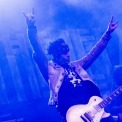 A blue tinted photo of a man with an electric guitar standing with his arms in the air while another guitar player plays behind him.