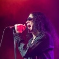 The side view of a man with long dark hair and sunglasses standing in purple light singing into a microphone.