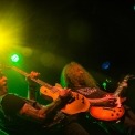 Two men wearing all black stand together playing electric guitars with yellow and green lights shining behind them.