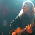 The side view of a man with long hair and glasses stands in a black button down shirt playing an acoustic guitar.