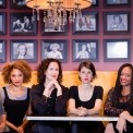 Six women in black clothing sitting at a table on a yellow couch with a red wall full of black and white pictures behind them.
