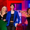 Three women in black, green and blue clothing. The woman in the middle is holding a microphone and the two women behind her are dancing in front of a red and blue background.