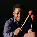 An African American man holds two drumsticks with red ends in front of a black background with his eyes closed.