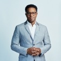 African American man wearing glasses in front of a bright blue background stands with his hands folded wearing a light gray suit jacket.