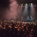 Six band members wearing black on stage playing instruments in front of a crowd of people all in front of a black background.