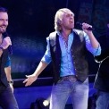 Three men stand singing into microphones wearing jeans, black vests, and blue button downs. The man on the right has on a white shirt and blue suit jacket.