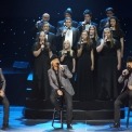 Three men sitting on stools singing into handheld microphones wearing black cowboy hats, black button down shirts, grey suit jackets, and black jeans stand in front of a choir of 12 college aged men and women standing on risers wearing long black dresses and tuxes singing into microphones. They are all in front of a black background with twinkling lights.