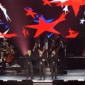 Three men standing wearing black button down shirts, thigh length black suit jackets, and black jeans sing into handheld microphones while standing in front of a choir of 12 college aged men and women standing on risers wearing long black dresses and tuxes singing into microphones. There is an orchestra of cello players to the left of them and violin players to the right. They are all in front of a background with red white and blue stars projected onto it.