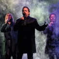 Three men stand in front of a black smoky background singing into microphones. They are wearing black button down shirts and thigh length black suit jackets.