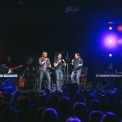 Three men stand on a stage in front of an audience wearing jeans and black button down shirts singing into handheld microphones with two keyboards on either side of them , a guitarist behind them and a black background with multicolored lights lining the left and right side of the stage.