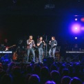 Three men stand on a stage in front of an audience wearing jeans and black button down shirts singing into handheld microphones with two keyboards on either side of them , a guitarist behind them and a black background with multicolored lights lining the left and right side of the stage.