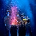 Three bald blue man standing in front of two drums pouring paint onto them in front of a blue and black background