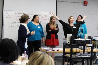 Image of five teachers standing at the front of a classroom and one teacher has her arms raised wide above her head.