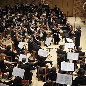 The Mariinsky Orchestra sits in a concert hall dressed in all black with music stands covered in sheet music. The conductor to the right is preparing to start their next piece.