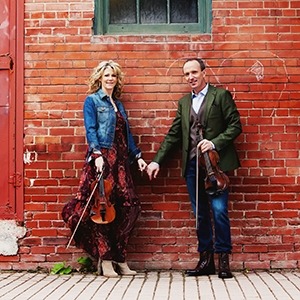 Image of the two performers from Celtic Celebration holding hands and their fiddles while standing in front of a brick wall.