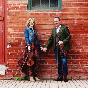 Image of the two performers from Celtic Celebration holding hands and their fiddles while standing in front of a brick wall.