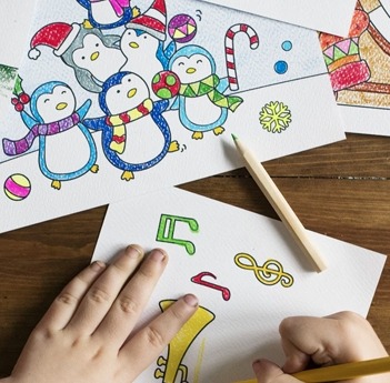 Image of two hands coloring a drawing with a yellow color pencil and an additional completed coloring pages on the table