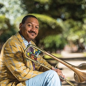 Delfeayo Marsalis wearing a gold jacket, sitting outside by some trees, holding his trombone.