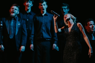 Image of Kristin Chenoweth on the Lied Center stage in a black dress singing powerfully into a microphone with the chorus standing behind her.