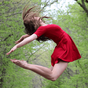 Image of female dancer in red dress. She is jumping in the air bending her arms and legs together behind her back. There is a backgound of forest trees behind her.