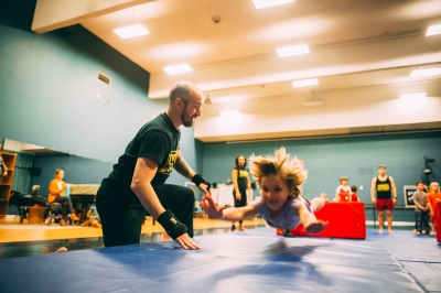 Image of a child flying through the air with her hands stretched forward about to land on a blue mat while a performer spots her during the STREB outreach program.