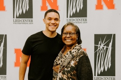 Image of Trevor Noah posing with a Lied Center donor in front of the white backdrop with red University of Nebraska logos and black Lied Center logos.